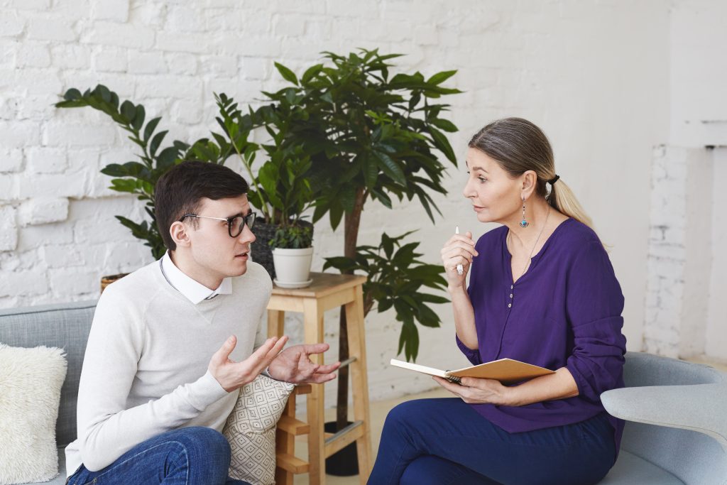 Talk to a Therapist Psychology, therapy, psychiatry, mental health and counseling concept. Candid shot of nervous self conscious young male in glasses telling middle aged female counselor about his problems at work